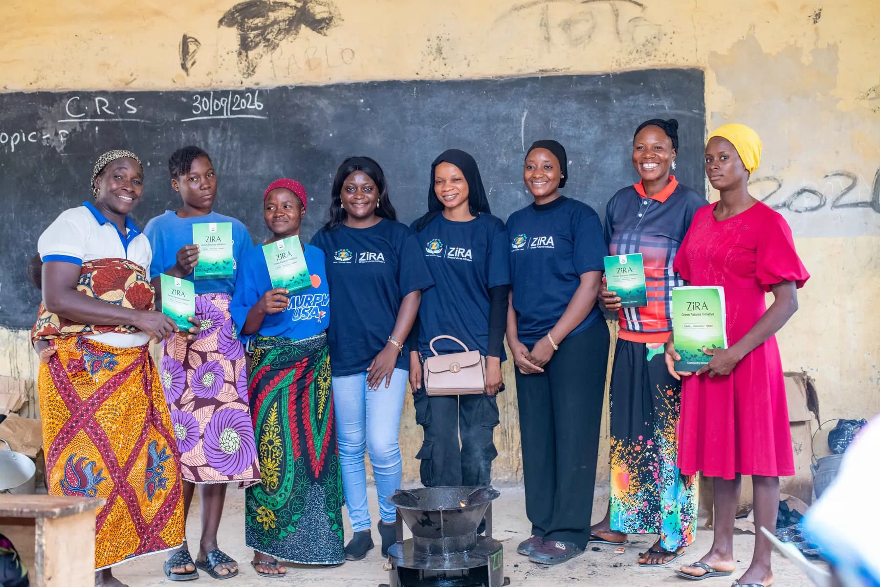 ZiRA team and Piko Village participants holding clean cooking workbooks beside a cookstove